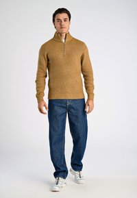 Ljusbrun ribbad stickad tröja med halvdragkedja i kragen, kombinerad med lös blå denimjeans och vita sportskor.