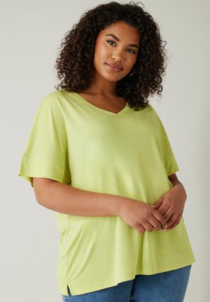 Vrouw met krullend haar, gekleed in een loszittend lichtgroen T-shirt met V-hals en korte mouwen, en een blauwe spijkerbroek, staand tegen een effen achtergrond.