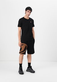 Black cotton t-shirt with a small red logo, paired with black shorts featuring a large brown graphic. Footwear is black athletic shoes.