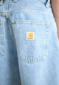 Shorts din denim de culoare albastră deschisă, cu o pată albă pătrată ce prezintă un logo galben și text roșu pe buzunarul din spate. Detalii evidente de cusături.