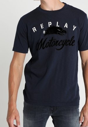 Mies, jolla on päällään merensininen T-paita, jossa on tekstit "Replay" ja "Motorcycle" sekä kotkan kuva rinnassa, yhdistettynä tummiin farkkuihin.