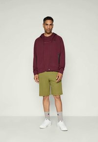 Bordeauxrode zip-up hoodie met voorzakken, gecombineerd met olijfgroene shorts en witte sneakers. Grijze sokken met branding zijn zichtbaar.