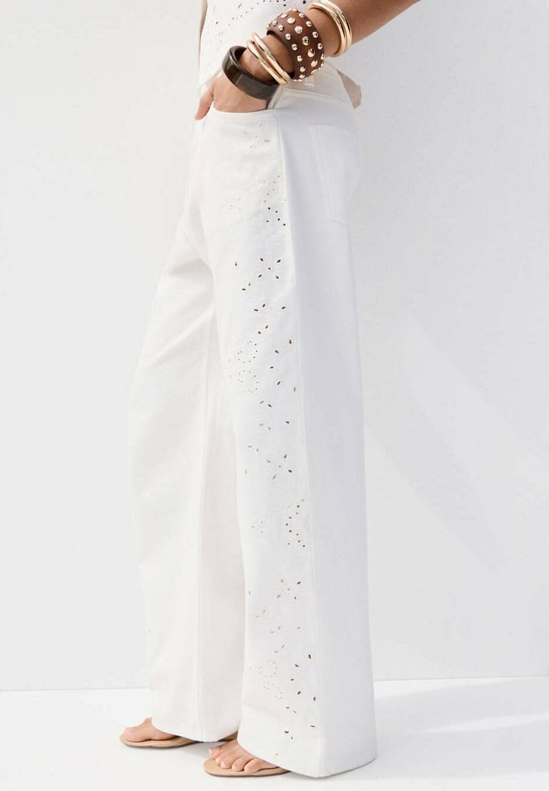 Persoon die witte wijd uitlopende broek draagt met bloemenuitgesneden details, hand in de zak en meerdere armbanden aan de pols, staand op blote voeten.