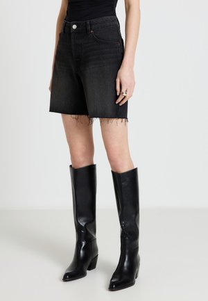 Donna che indossa pantaloncini di jeans neri a vita alta con orlo sfrangiato e stivali di pelle neri alti fino al ginocchio, in piedi contro uno sfondo semplice.