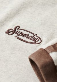 Lysegrå bomuldsstof med brun "Superdry" logo broderi. Har en kontrasterende brun ribstrik ved ærmet.