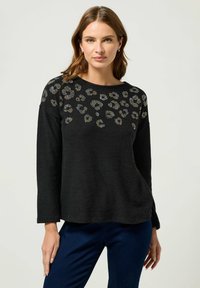 Schwarzer Pullover mit langen Ärmeln, der ein strukturiertes Tiermuster und goldene sowie silberne Nieten entlang des Ausschnitts aufweist. Weiches Material und entspannter Schnitt.