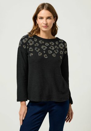 Schwarzer Pullover mit langen Ärmeln, der ein strukturiertes Tiermuster und goldene sowie silberne Nieten entlang des Ausschnitts aufweist. Weiches Material und entspannter Schnitt.