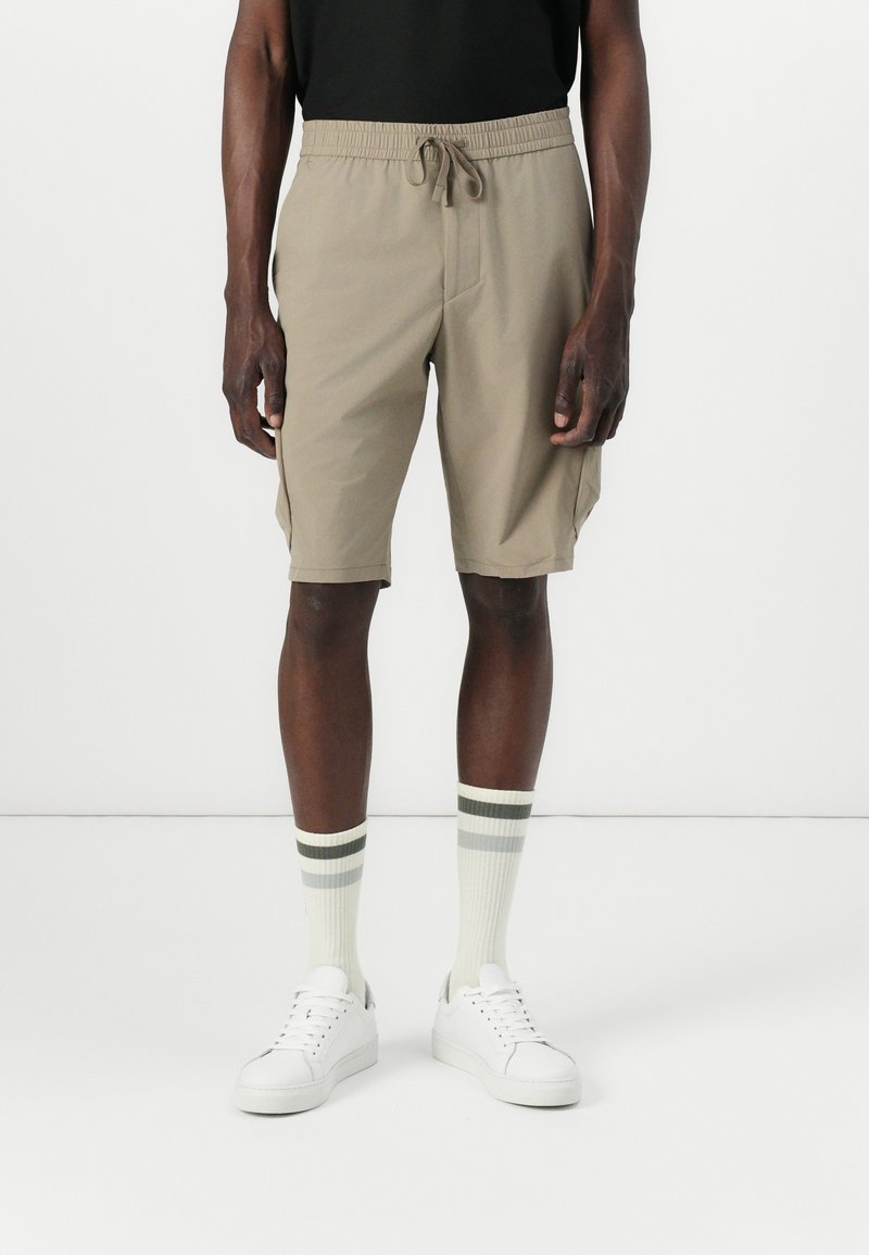Beige shorts med elastisk midjebånd og snøring. Laget av lett materiale, med sidelommer og en avslappet passform. Hvite sneakers og stripe strømper.