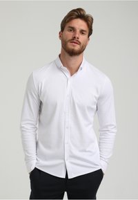Camisa blanca de manga larga con botones, con cuello extendido, hecha de tejido suave, que presenta un corte regular y un sutil detalle de costuras.