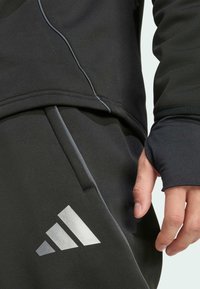 Der schwarze sportliche Sweatshirt und die Jogginghose verfügen über einen glatten Stoff, eine enganliegende Passform, reflektierende Akzente und ein auffälliges silbernes Logo am Bein.