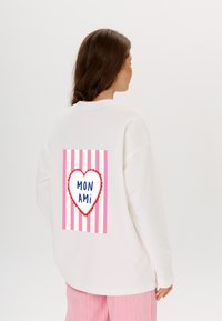 Weiße oversized Sweatshirt mit einem pinken und roten Herzdesign auf dem Rücken, das den Text "MON AMI" innerhalb eines weißen Herzens und einem gestreiften Hintergrund zeigt.