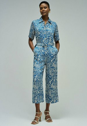 Mujer de pie con las manos en los bolsillos, vestida con un mono de estampado floral azul y blanco y tacones beige con tiras sobre un fondo gris.