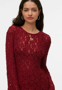 Bordowy top z kwiatowej koronki z długim rękawem, z teksturowanym wzorem i okrągłym dekoltem, noszony na cielistym podkoszulku. Akcent w postaci złotego naszyjnika.