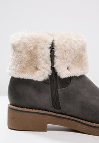 Bottine en simili cuir gris avec un revers en fausse fourrure crème, fermeture éclair sur le côté et semelle texturée marron, conçue pour le confort et la chaleur.