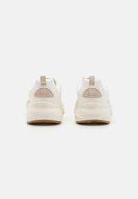 Bílé sportovní boty s hladkým povrchem, síťovinovými detaily a světlou béžovou gumovou podrážkou. Mají zadní poutko pro snadné nazouvání.