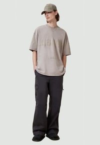 Beige oversized t-shirt with embossed branding, paired with black cargo pants featuring pockets and black slip-on shoes. Gray cap worn.