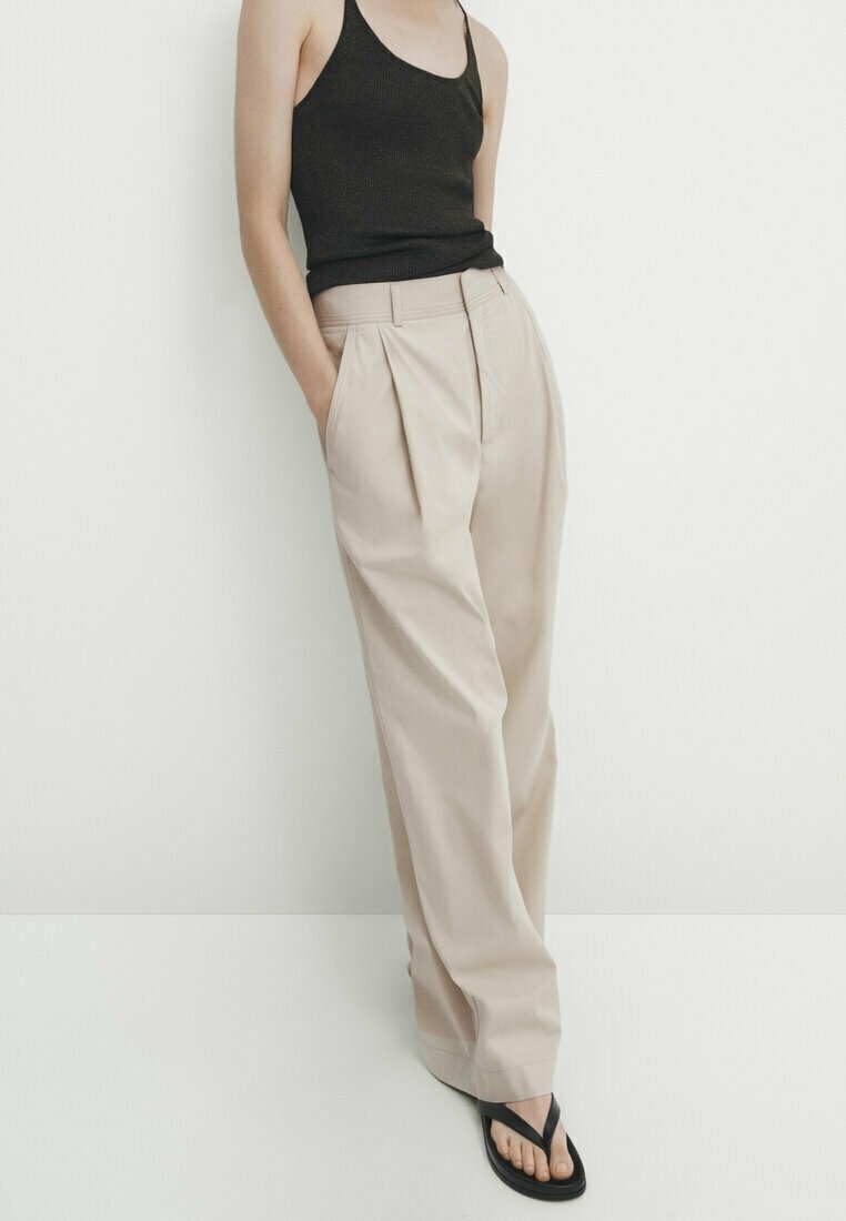 Personne portant un pantalon plissé beige taille haute, un haut noir sans manches et des tongs noires, debout les mains dans les poches sur fond blanc.