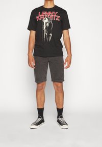 Persona con camiseta gráfica negra de Lenny Kravitz, pantalones cortos gris oscuro, calcetas negras y zapatillas bajas negras, de pie contra un fondo blanco.