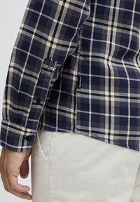 Cămașă cu guler tip button-down, în carouri bleumarin și crem, cu mâneci lungi și tiv curbat. Include nasturi negri și un etichetă cu logo pe lateral.