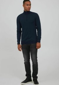 Jersey azul marino con cuello con cremallera y mangas largas, diseño ajustado y puños de canalé, combinado con jeans gris oscuro y zapatillas negras.