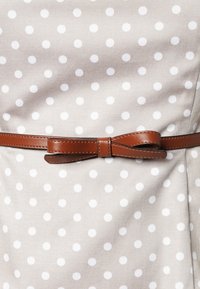 Tessuto beige con pois bianchi, rifinito con una cintura in pelle marrone con un dettaglio a fiocco, che enfatizza il contrasto di texture e pattern.