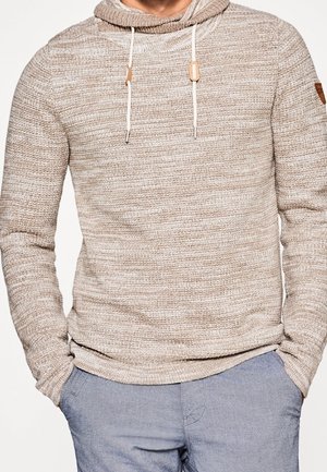 Mann trägt beige gestrickten Hoodie mit Kordelzügen und hellblaue Hose, Hände in den Taschen, steht vor einfarbigem Hintergrund.