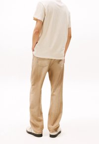 Beige ylin tee-paita, pyöreä kaula-aukko, yhdistettynä vaaleanruskeisiin, löysiin, teksturoituihin housuihin ja valkoisiin lenkkareihin, nähtynä takaa.