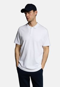 Biała koszulka polo z krótkim rękawem, z klasycznym kołnierzykiem i trzema guzikami. Model nosi ciemnogranatową czapkę. Czysty design, casualowy krój.