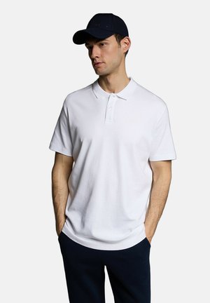 Biała koszulka polo z krótkim rękawem, z klasycznym kołnierzykiem i trzema guzikami. Model nosi ciemnogranatową czapkę. Czysty design, casualowy krój.