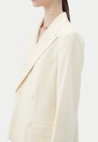 Blazer croisé couleur beige crème, doté d'un col à revers pointu, de deux boutons et d'une poche poitrine unique avec des détails de coutures visibles.