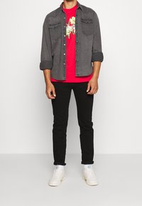 Muž v tmavě šedé košili na knoflíky přes červené květinové tričko, černých kalhotách, bílých ponožkách a bílých teniskách stojící proti jednobarevnému pozadí.