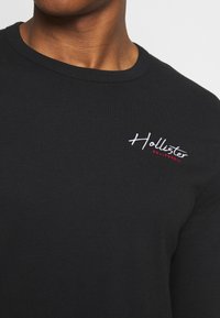 Svart långärmad t-shirt i bomull. Har rundad halsringning och broderad "Hollister"-logotyp i vitt med röda detaljer. Slät textur.