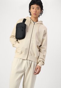 Beige Zip-up-Hoodie aus weichem Stoff mit einer Fronttasche, getragen über hellen, lässigen Hosen. Eine schwarze Umhängetasche ist über die Schulter geschlungen.