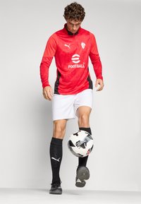 Rød langermet fotballtrøye med svarte detaljer, hvite shorts og svarte sokker. Har Puma-logo og teksturert mønster. Spiller som balanserer en fotball.