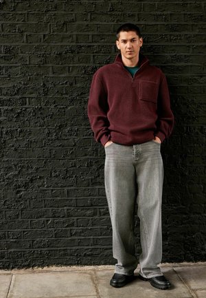 Bordeaux fleece trui met een ritssluiting en borstzak, gecombineerd met losse grijze jeans en zwarte gestructureerde schoenen, tegen een zwarte bakstenen muur.