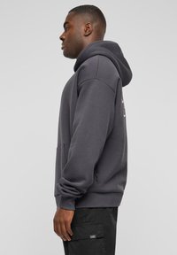 Mørkegrå hettegenser laget av myk bomullsblanding, med en romslig kengurulomme, ribbede mansjetter, og et trykt logo på ryggen.