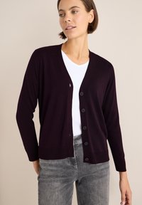 Dunkellila Strickjacke mit V-Ausschnitt, Knopfverschluss und langen Ärmeln. Aus weichem Stoff mit gerippten Bündchen und Saum. Über einem weißen T-Shirt getragen.