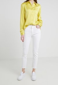 Camisa amarilla de satén con botones y un bolsillo en el pecho, combinada con jeans ajustados blancos y zapatillas blancas. Textura suave y diseño informal.