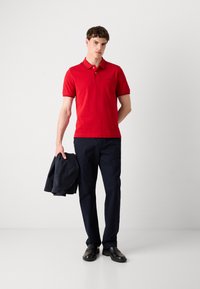 Crvena polo majica s kratkim rukavima, ovratnikom i tamnim obrubom, u kombinaciji s tamnoplavim hlačama i crnim cipelama. Model drži tamni sako.