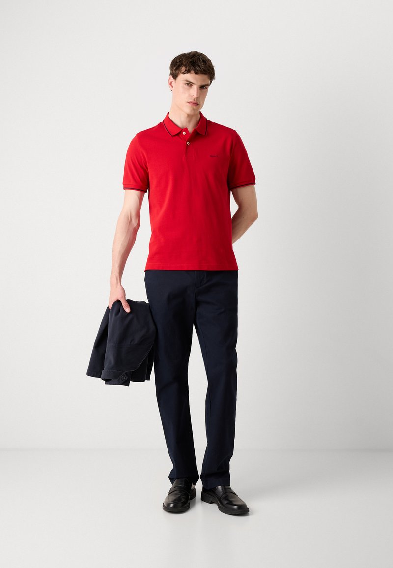 Polo rouge à manches courtes, col et bordure foncée, associé à un pantalon marine et des chaussures noires. Modèle tenant une veste sombre.