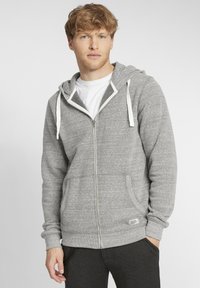 Siva zip-up dukserica s bijelim vezicama, kengurskim džepovima, rebrastim manžetama i tkanom etiketom na rubu, uparena s bijelom majicom ispod.