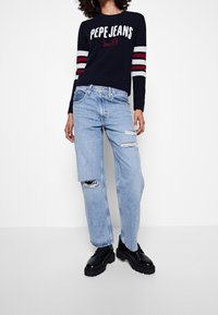 Pull marin avec rayures blanches et rouges, affichant le texte « PEPE JEANS ». Jean bleu clair déchiré avec ourlets bruts. Chaussures noires à plateforme.