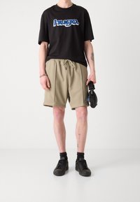 Musta puuvillainen t-paita, jossa sininen logo, beigefarkut nyörillä ja mustat tennarit. Tasainen pinta, rento muotoilu, vähäiset yksityiskohdat.