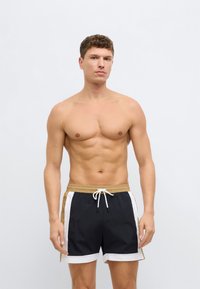 Čierne plavecké šortky so zlatými akcentmi, bielymi bočnými pruhmi, nastaviteľným sťahovacím pásom, ľahkým materiálom a dĺžkou po polovicu stehna.