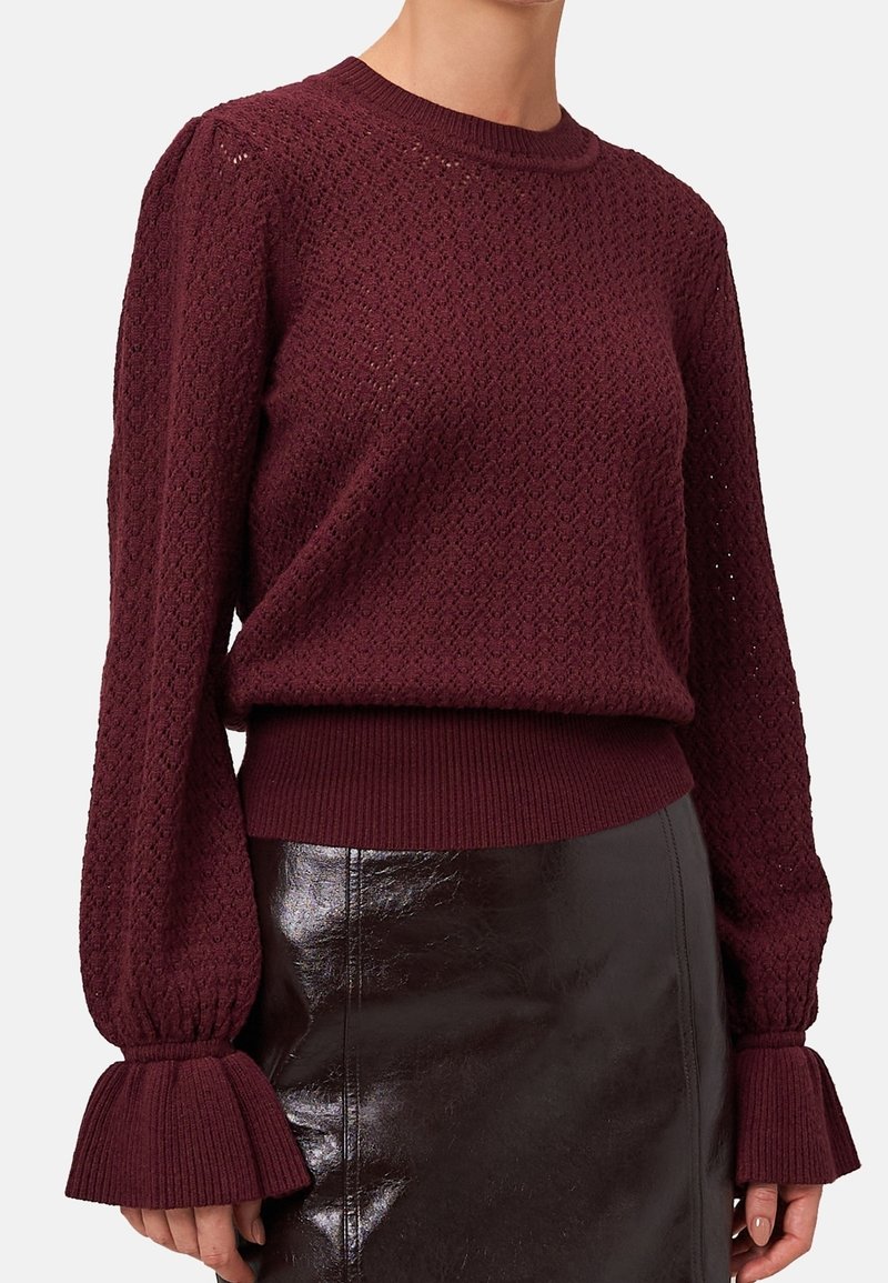 Femme portant un pull en maille texturée bordeaux avec des manches bouffantes et des poignets côtelés évasés, associé à une jupe en cuir marron foncé.