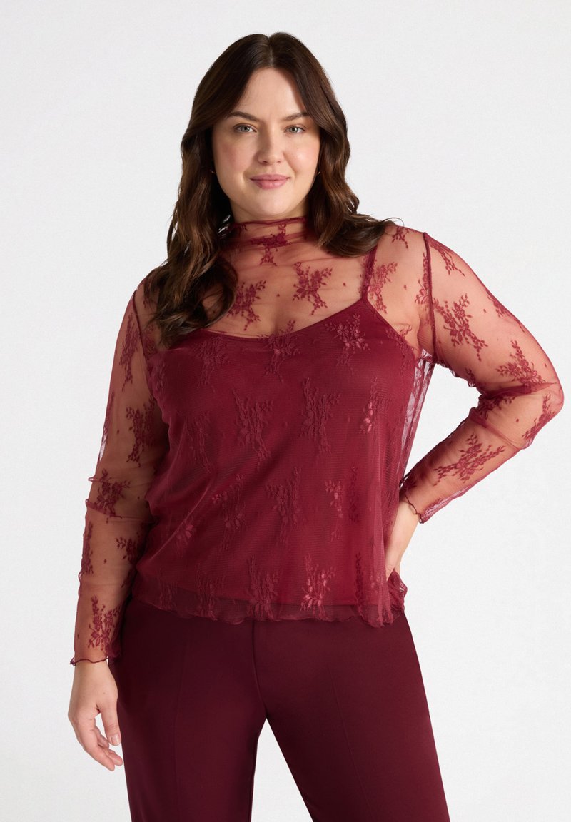 Burgundi pitsile top, joka on varustettu pitkillä läpinäkyvillä hihoilla, korkealla kauluksella ja kukkakuviolla, kerroksittain tiukan tankopaidan päällä.