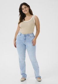 Débardeur beige à côtes avec des empiècements blancs, associé à un jean taille haute bleu clair. Le modèle porte des chaussures slip-on beiges. Textures lisses, coupe décontractée.