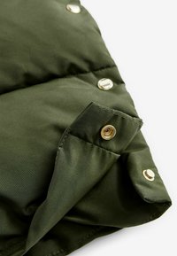 Zelena puffer jakna s metalnim kopčama, koja se ističe kvadratnim uzorkom i glatkom tkaninom, s naglaskom na otvorenoj džepu.