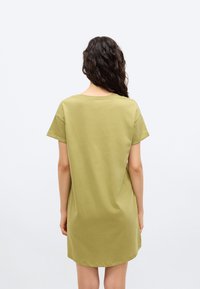 Γυναίκα με μακριά σγουρά σκοτεινά μαλλιά, ντυμένη με φαρδιά ελιάς πράσινη κοντομάνικη φόρεμα, όρθια με την πλάτη προς το φακό, μπροστά σε ένα απλό φωτεινό φόντο.