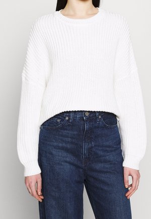 Personne portant un pull blanc à manches longues côtelé, rentré dans un jean taille haute bleu foncé, sur un fond uni.